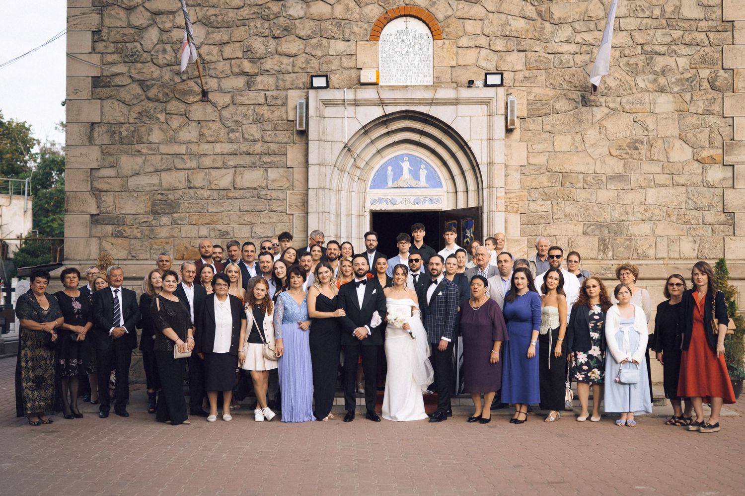 Large wedding party group photo outside a stone church with bride and groom in the center surrounded by guests.