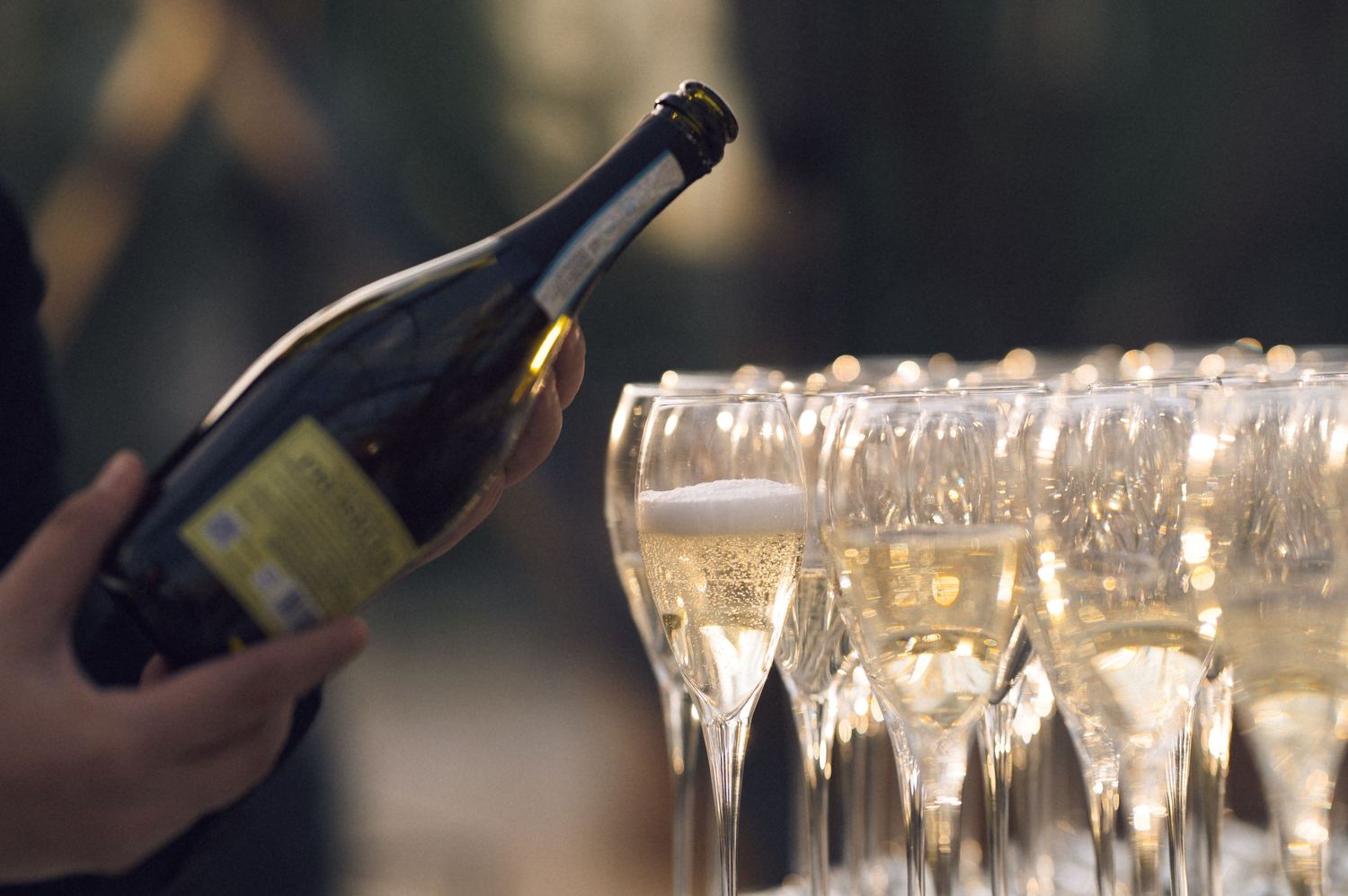 Person pouring champagne from a bottle into a row of elegant flute glasses at a celebration event.