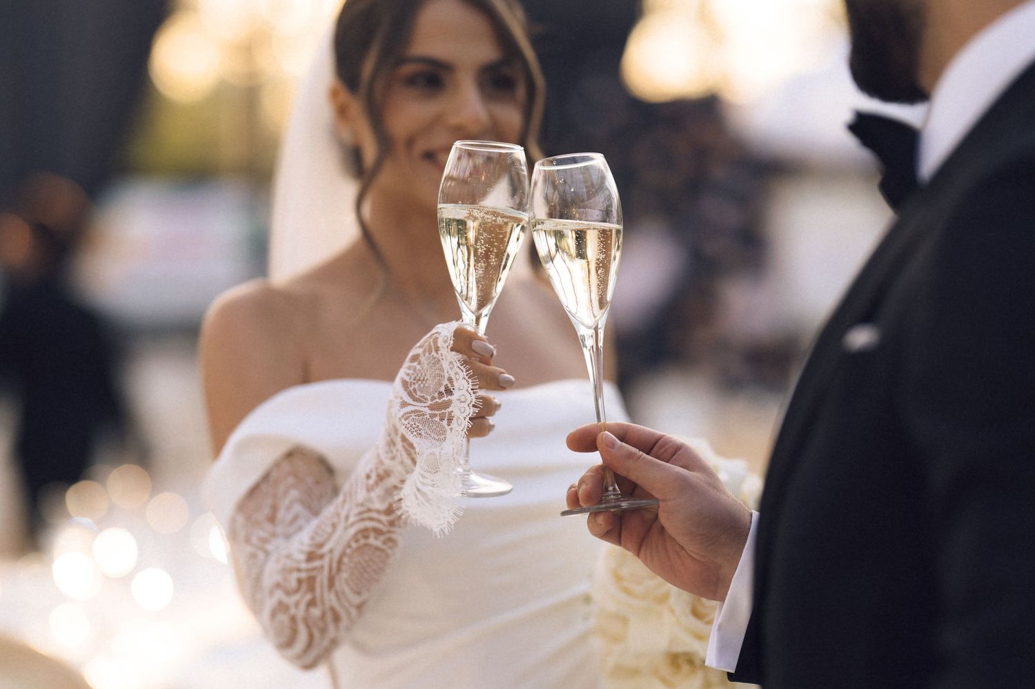 Bride and groom clinking champagne glasses at wedding reception, bride wearing elegant lace off-shoulder white gown.