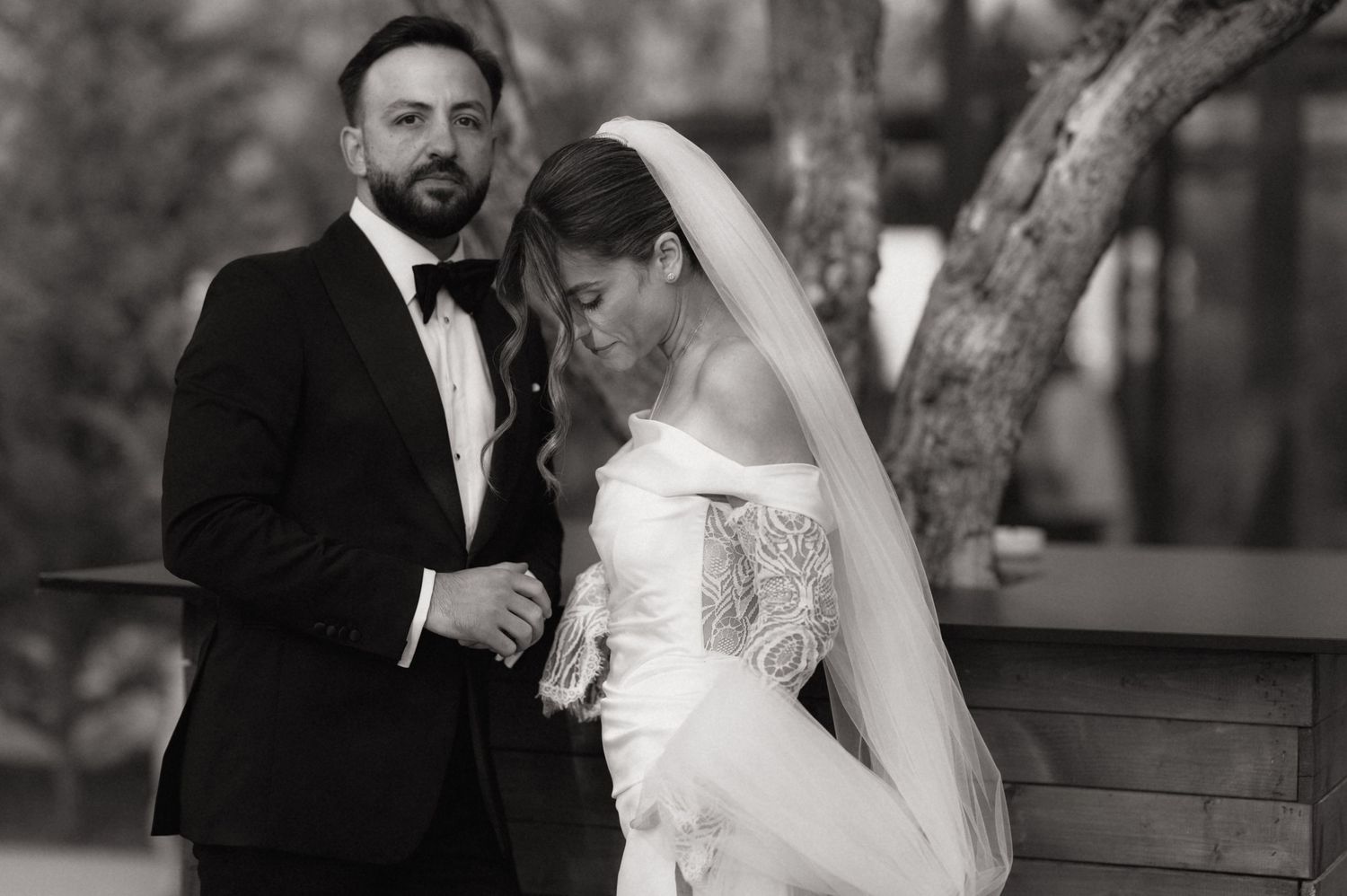 Black and white wedding photo of bride in lace gown with veil and groom in tuxedo standing outdoors.