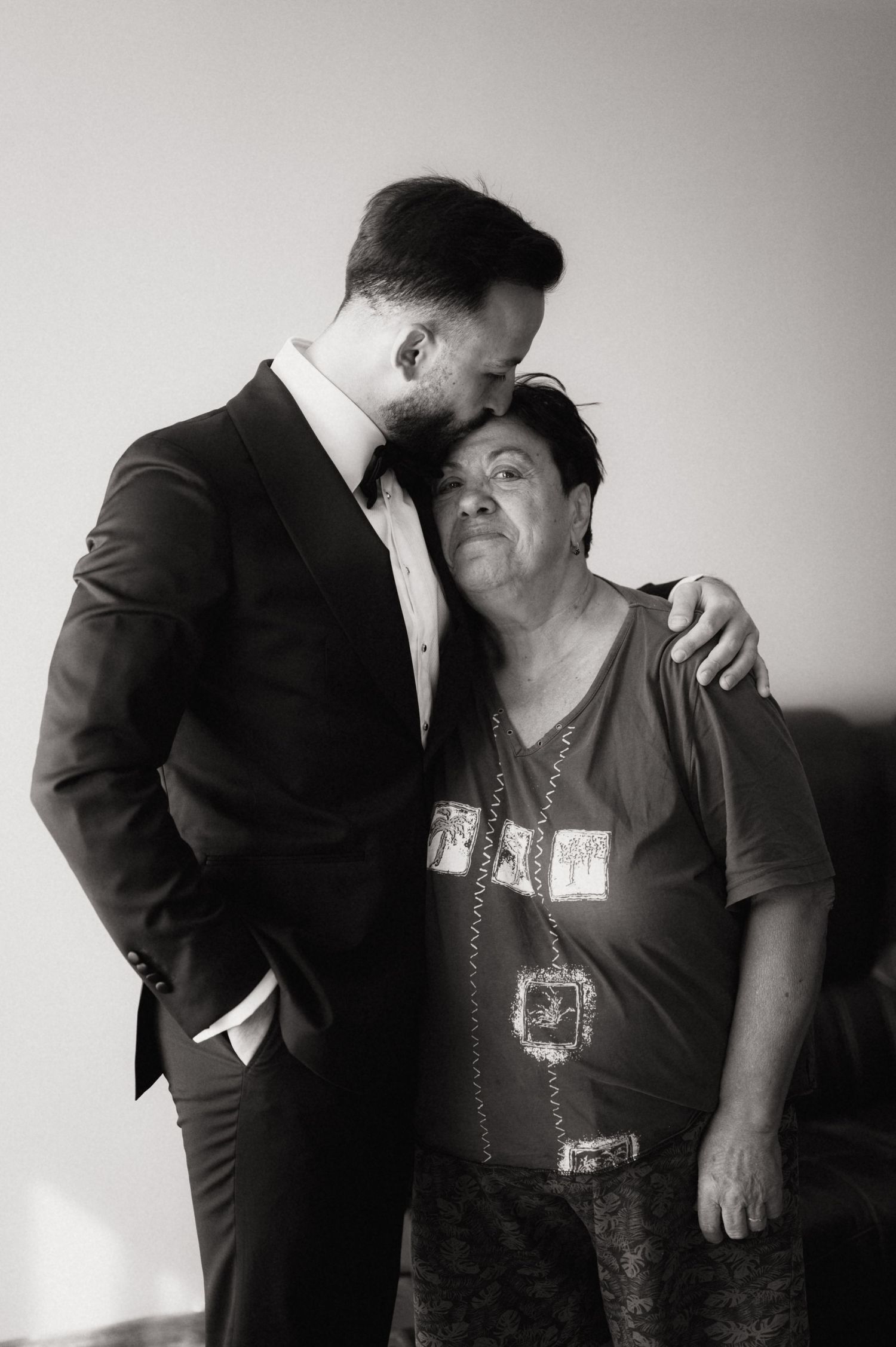 Black and white photo of a man in a suit tenderly kissing an older woman on the cheek, sharing a warm embrace.