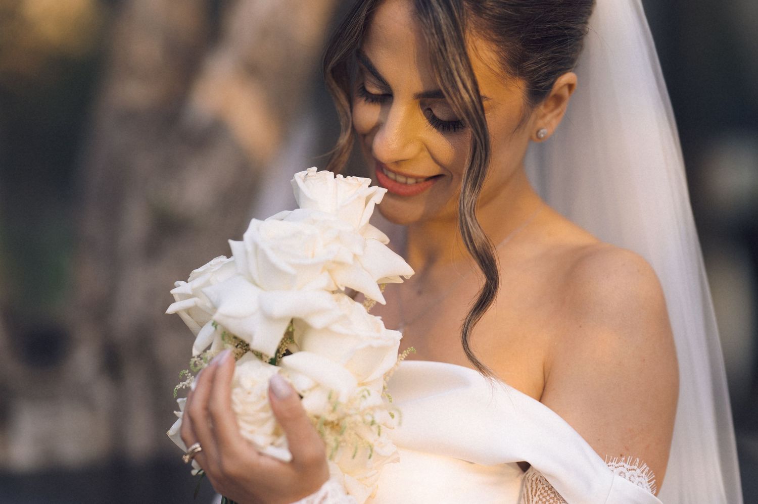 Bride in white off-shoulder dress smelling white rose bouquet, wearing veil, smiling softly outdoors.