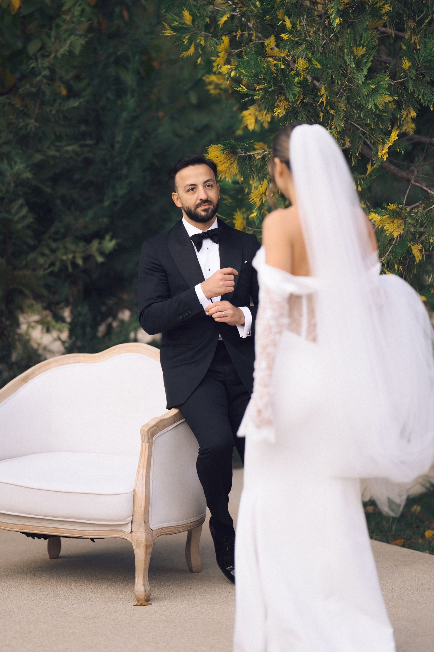 Groom in black tuxedo gazes at bride in white lace wedding dress and veil during outdoor first look.