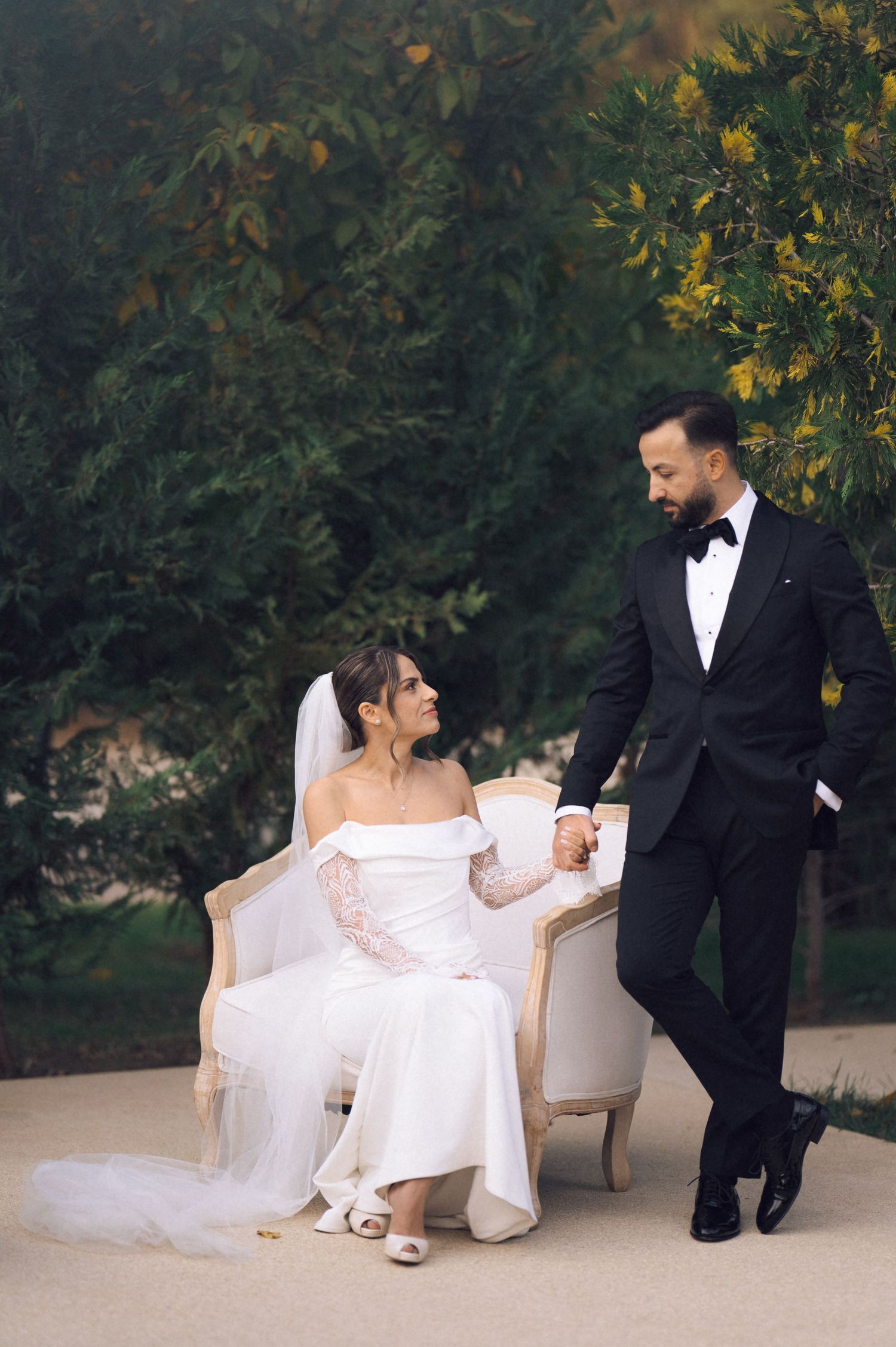 Bride in off-shoulder gown sits on vintage chair holding groom's hand in black tuxedo, posing outdoors amid lush greenery.