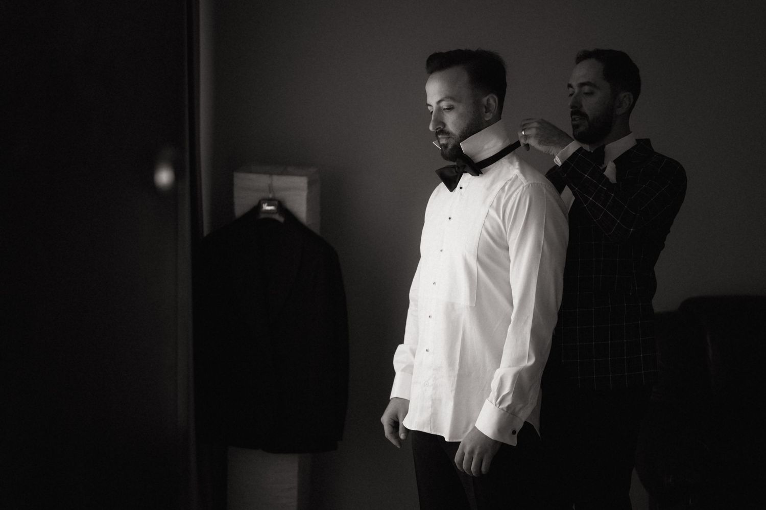 Black and white photo of a groom in a white shirt getting his bow tie adjusted by a groomsman before the wedding.