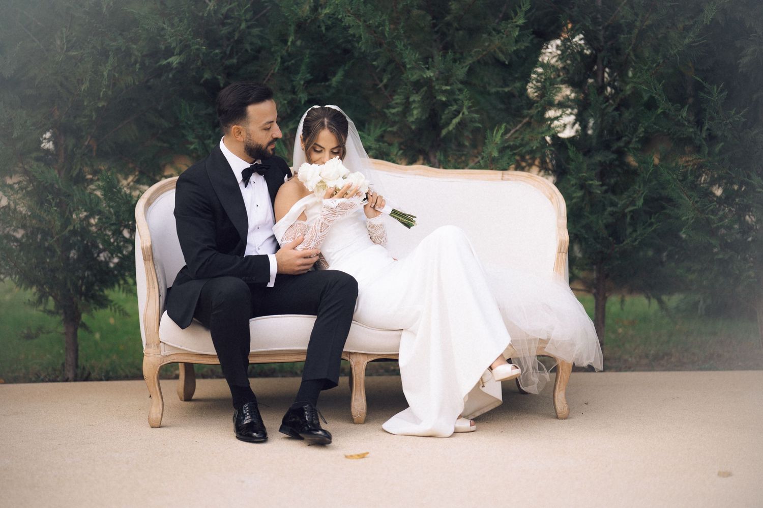 Bride and groom sitting together on an elegant white sofa outdoors, sharing a romantic moment on their wedding day.