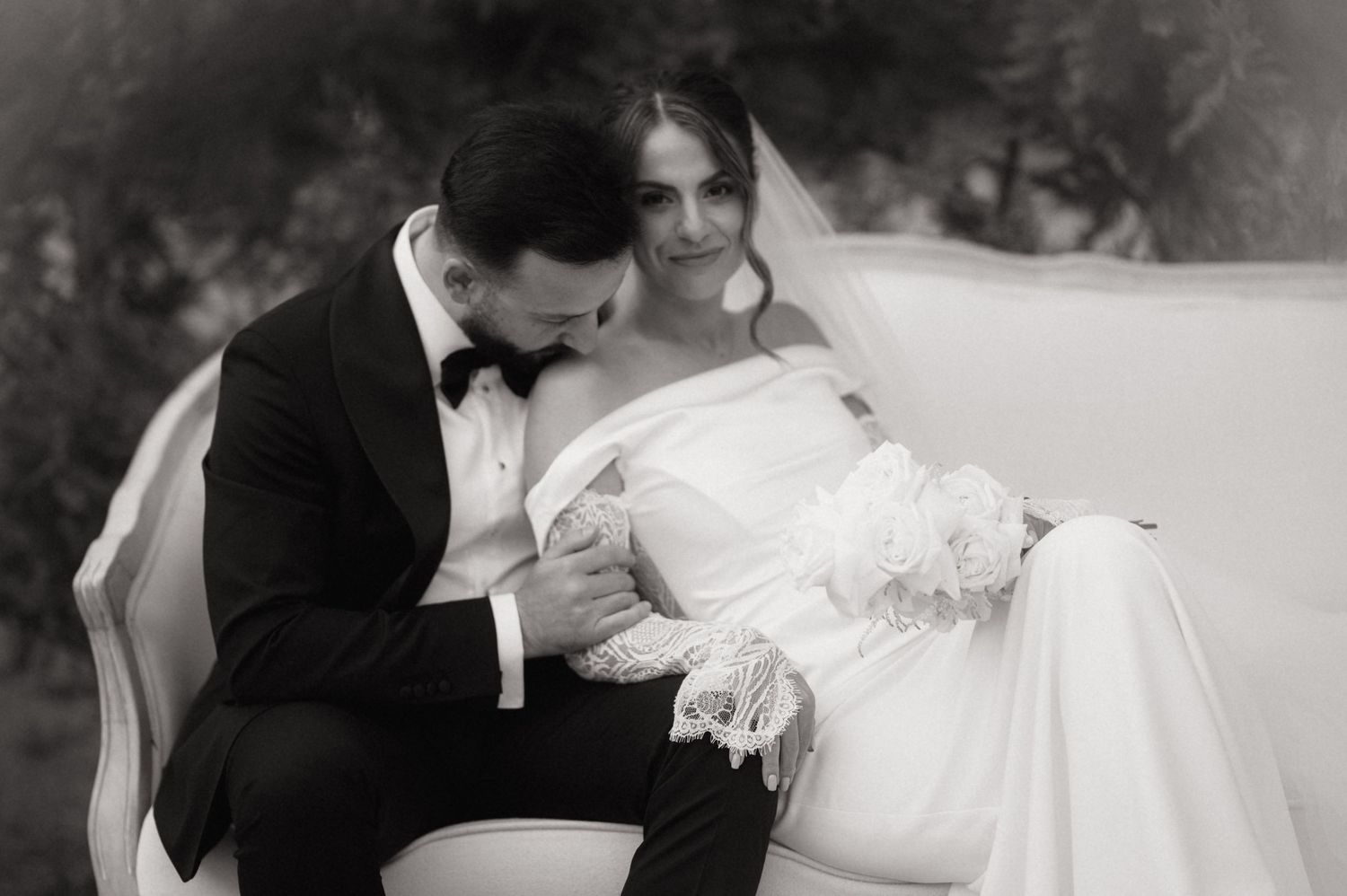 Romantic black and white wedding photo of groom in tuxedo tenderly kissing brides hand as she reclines on sofa.