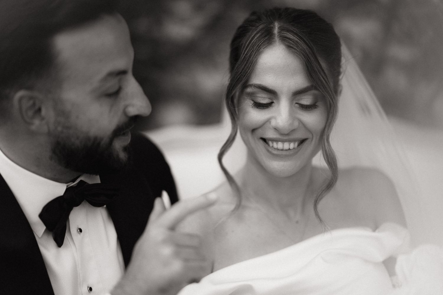 Black and white wedding photo of smiling bride in veil with groom in tuxedo and bow tie looking down tenderly.