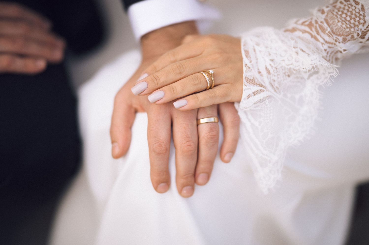 Close-up of newlywed couple's hands displaying matching gold wedding rings, bride wearing elegant lace wedding dress.