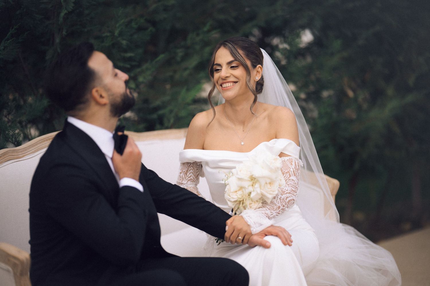 Bride in white off-shoulder gown holding bouquet sits with groom in black tuxedo on elegant outdoor sofa.