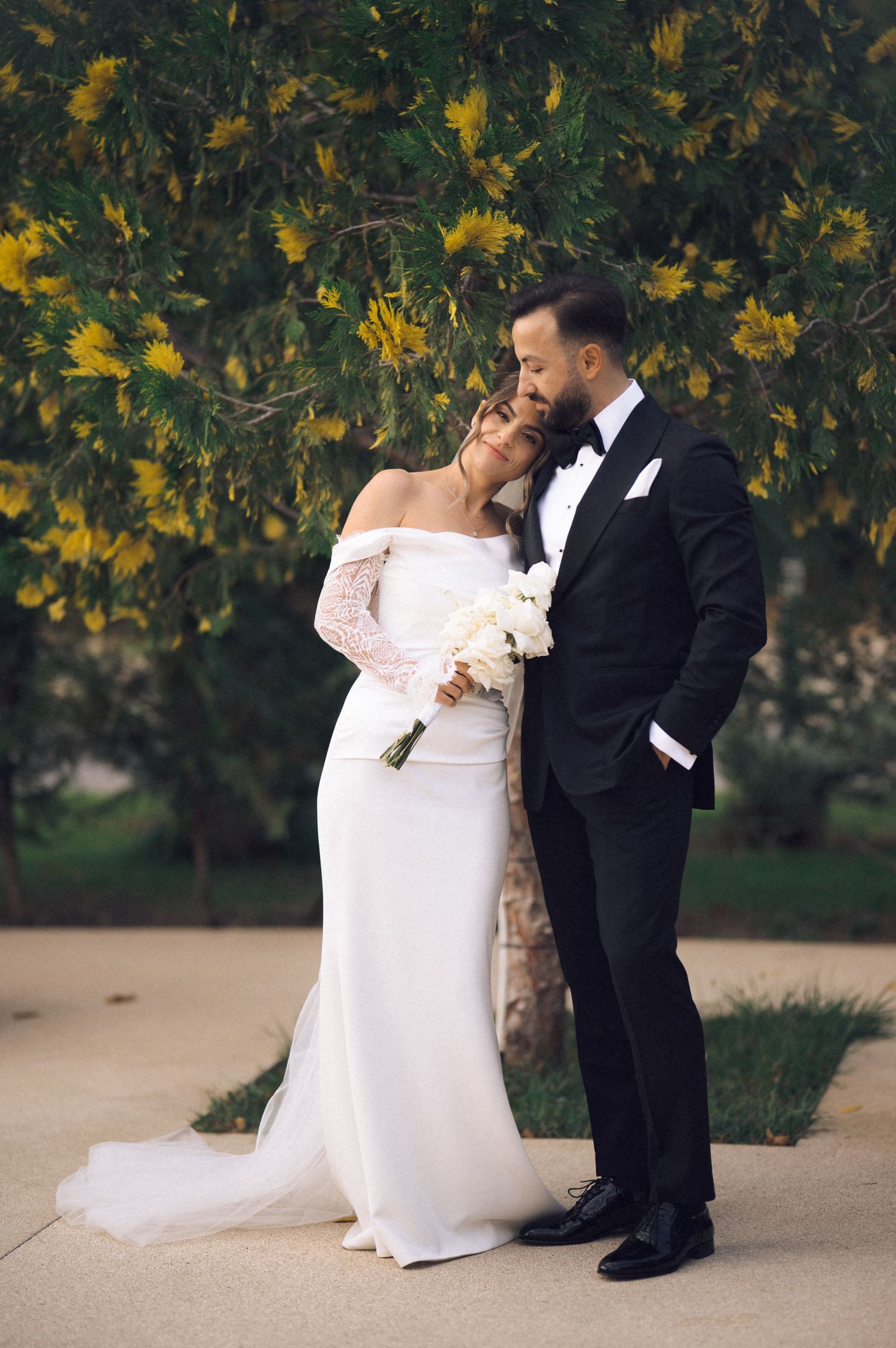 Bride and groom embracing before yellow flowering tree, she in white off-shoulder gown holding bouquet, he in black tuxedo.