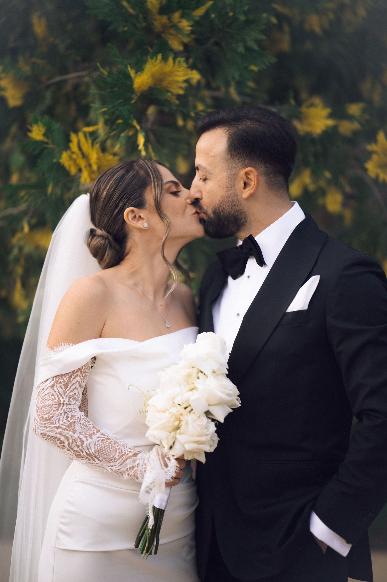 Bride and groom sharing a romantic kiss, she holds white bouquet in lace gloves, he wears black tuxedo, outdoors.
