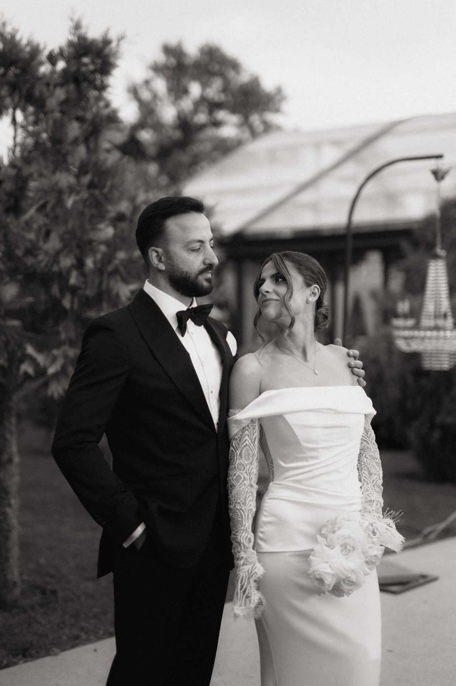 Black and white wedding photo of bride in off-shoulder lace gown and groom in tuxedo posing outdoors.