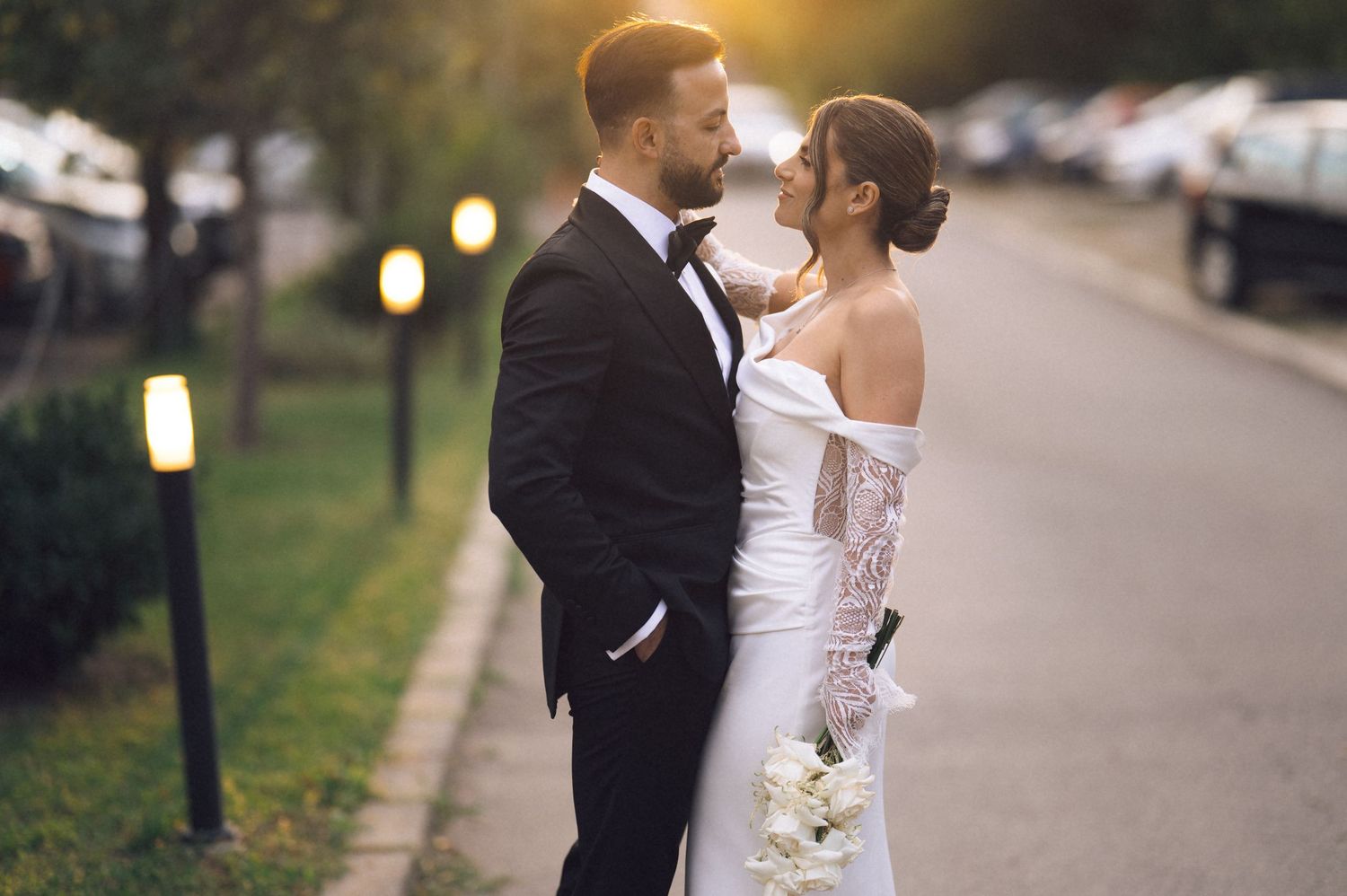 Bride and groom share a romantic moment on a tree-lined path at sunset, she in a white gown, he in a black tuxedo.