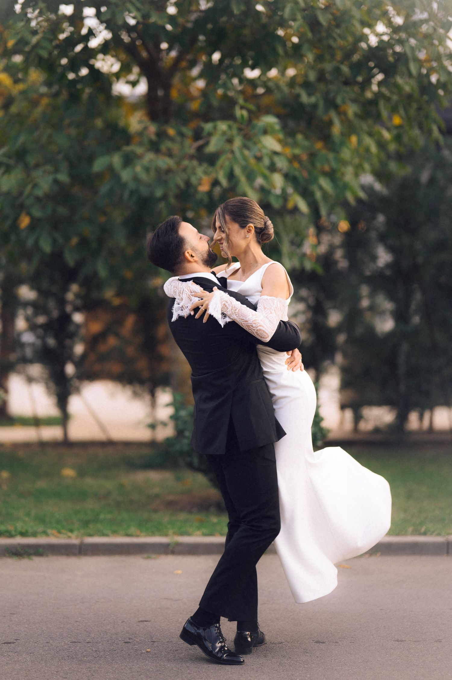 Bride and groom embrace outdoors, dancing joyfully in wedding attire with lush green trees in the background.