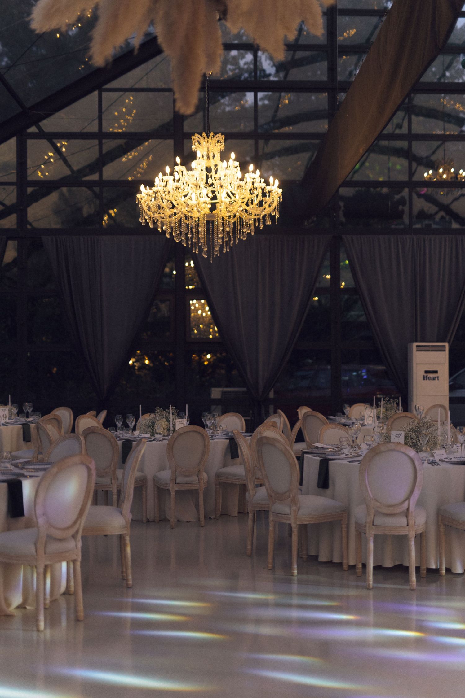 Elegant wedding reception venue with crystal chandelier, round tables, ivory chairs, and a white dance floor under a glass ceiling.