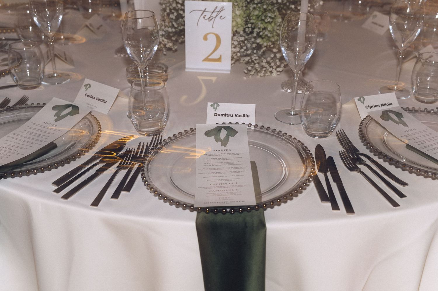 Elegant wedding reception table setting with charger plates, black napkins, glassware, and a gold table number 2 centerpiece.