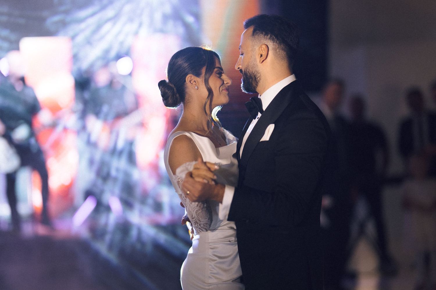 Bride and groom share their first dance at elegant wedding reception with sparkling lights in background.