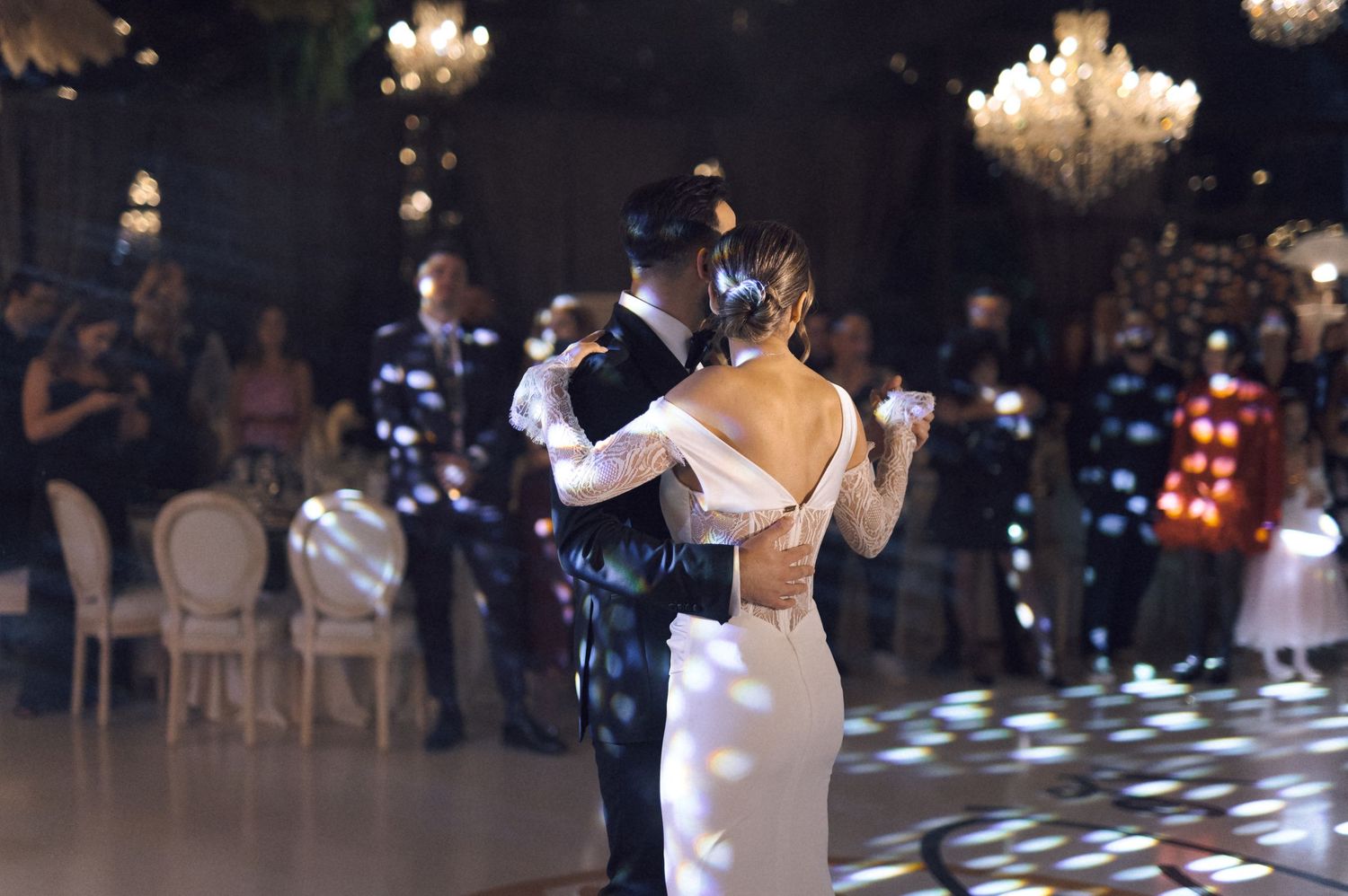 Couple sharing their first dance at an elegant wedding reception with guests watching and chandeliers glowing in the background.