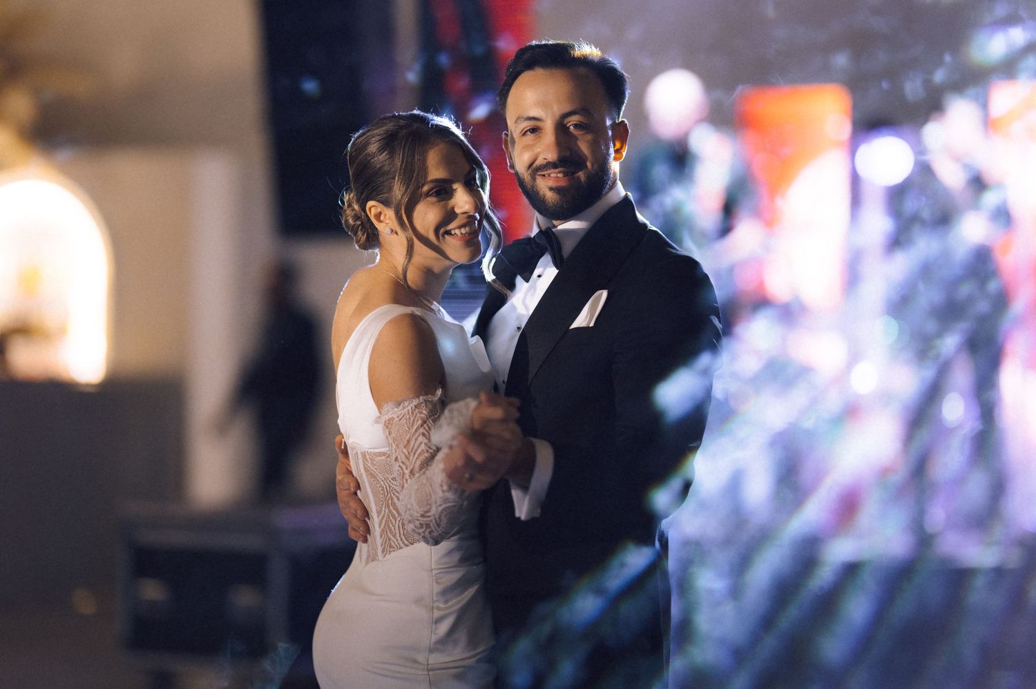 Elegant couple sharing first dance at wedding reception, bride in white gown and groom in black tuxedo, romantic evening lighting.
