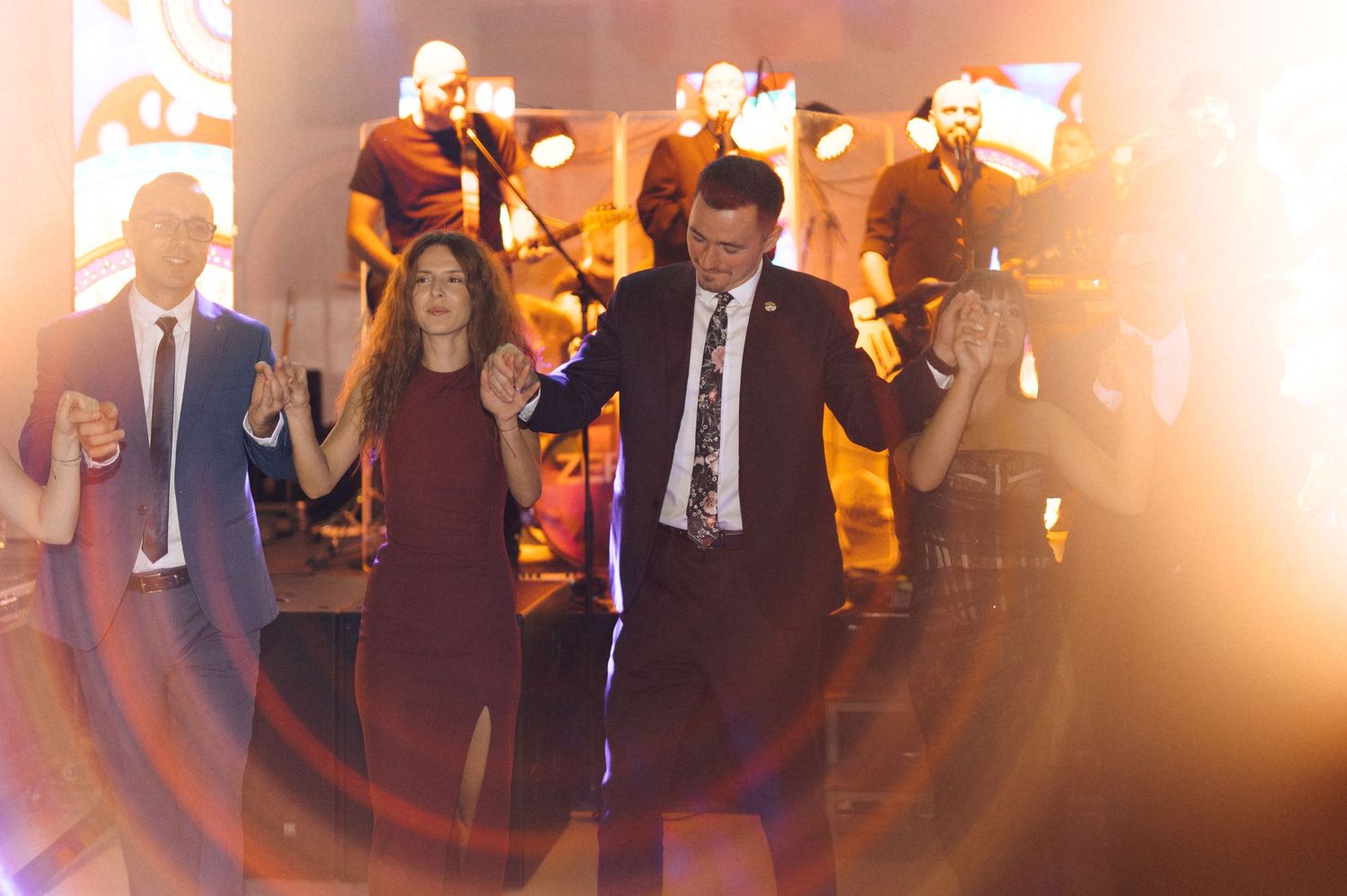 Three people hold hands and dance on a stage with a live band performing behind them at an elegant event.