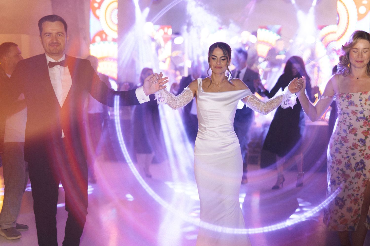 Bride in white gown dancing with guests at elegant wedding reception with purple lighting.