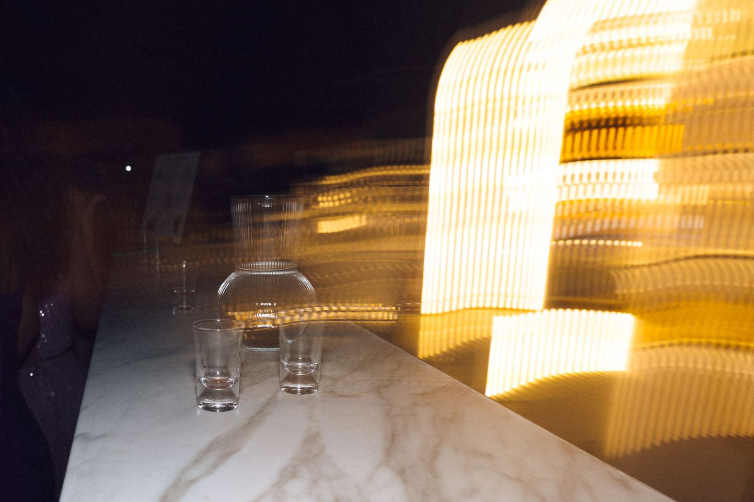 Blurred long-exposure photo of glowing amber lights beside glass cups on a marble surface at night.