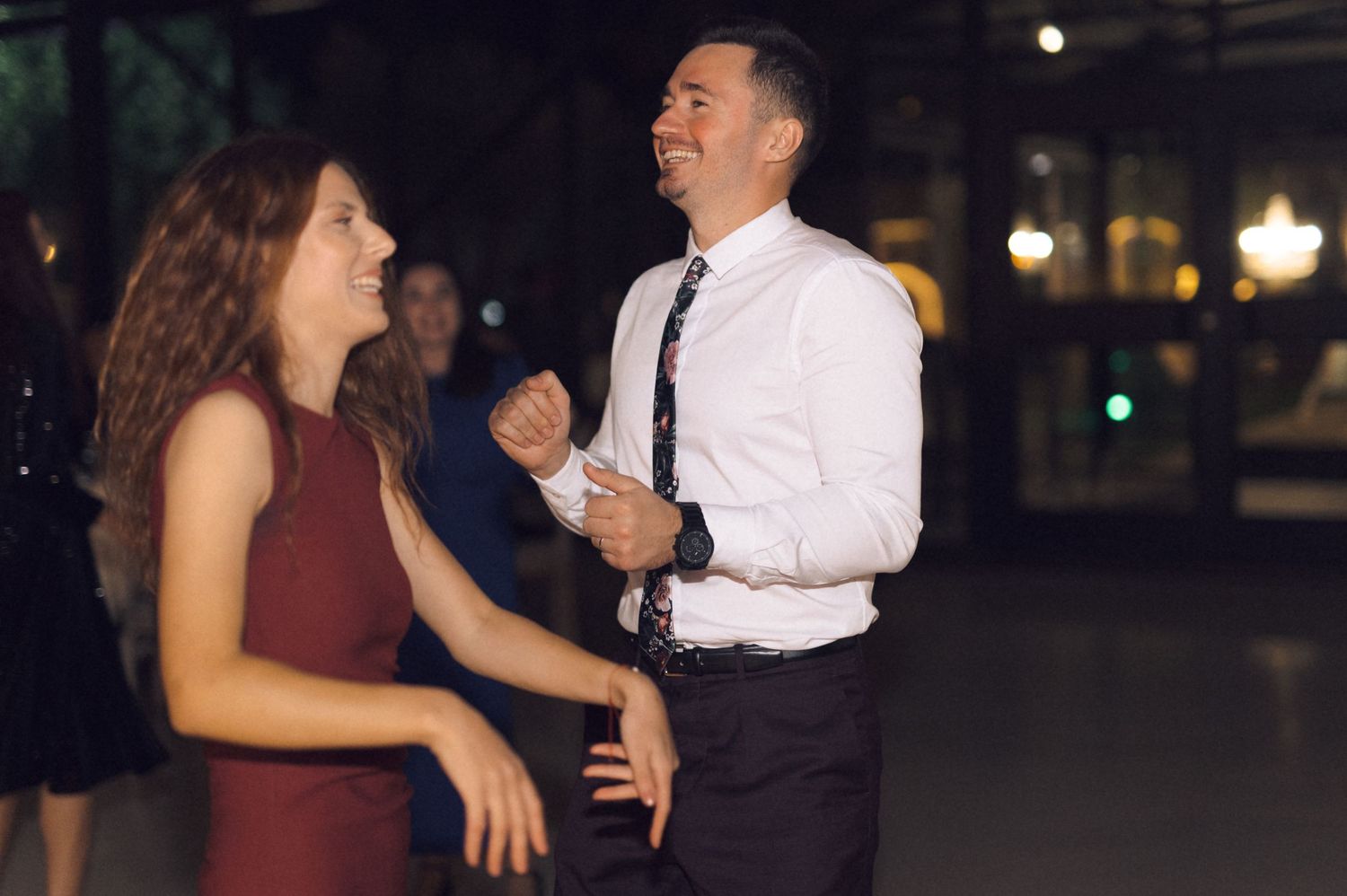 A man in a white shirt and tie laughs while dancing with a woman in a burgundy dress at an evening event.