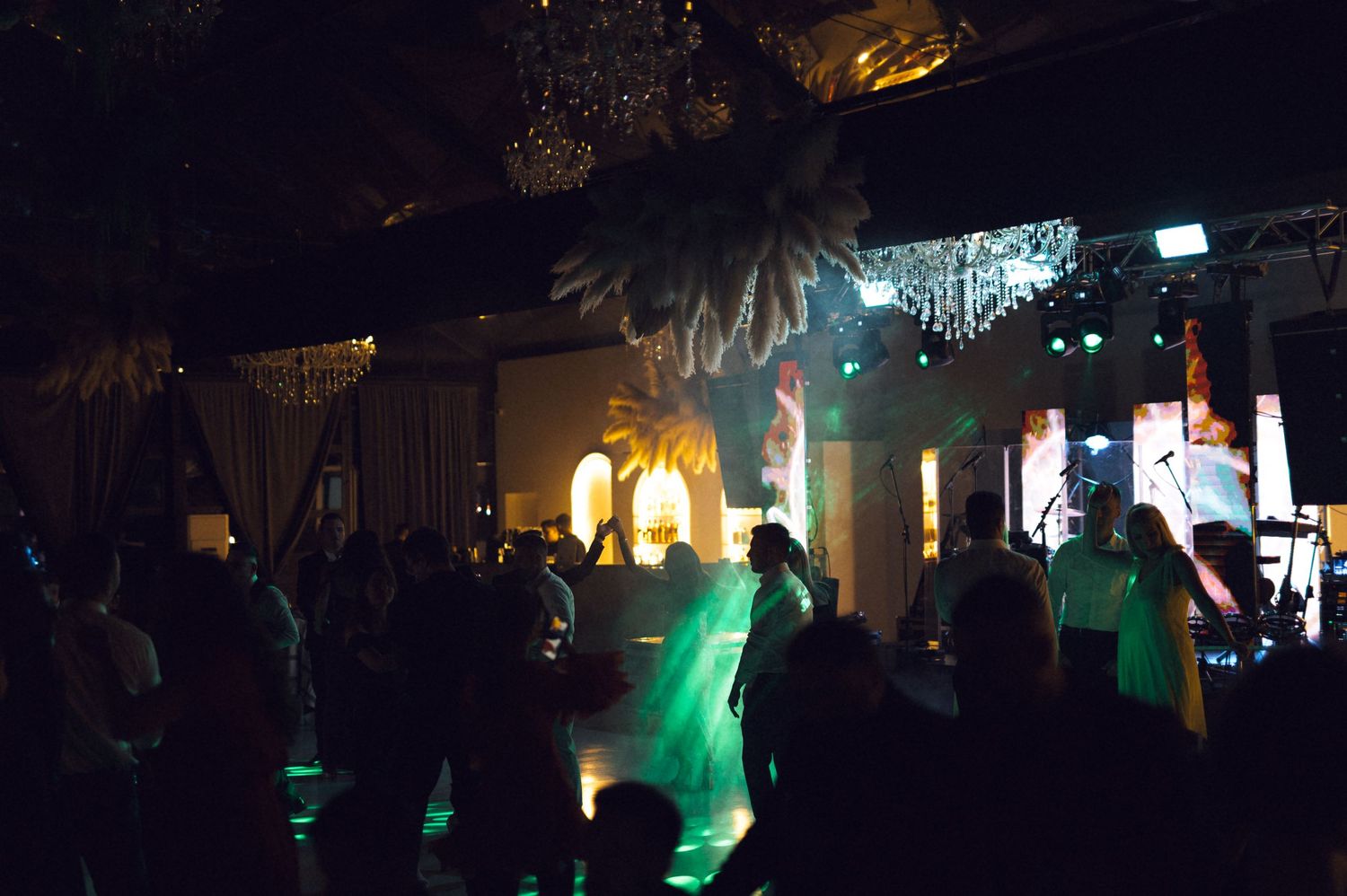 Crowded nightclub dance floor with green stage lighting, palm tree decor, and performers on stage in a dark venue.