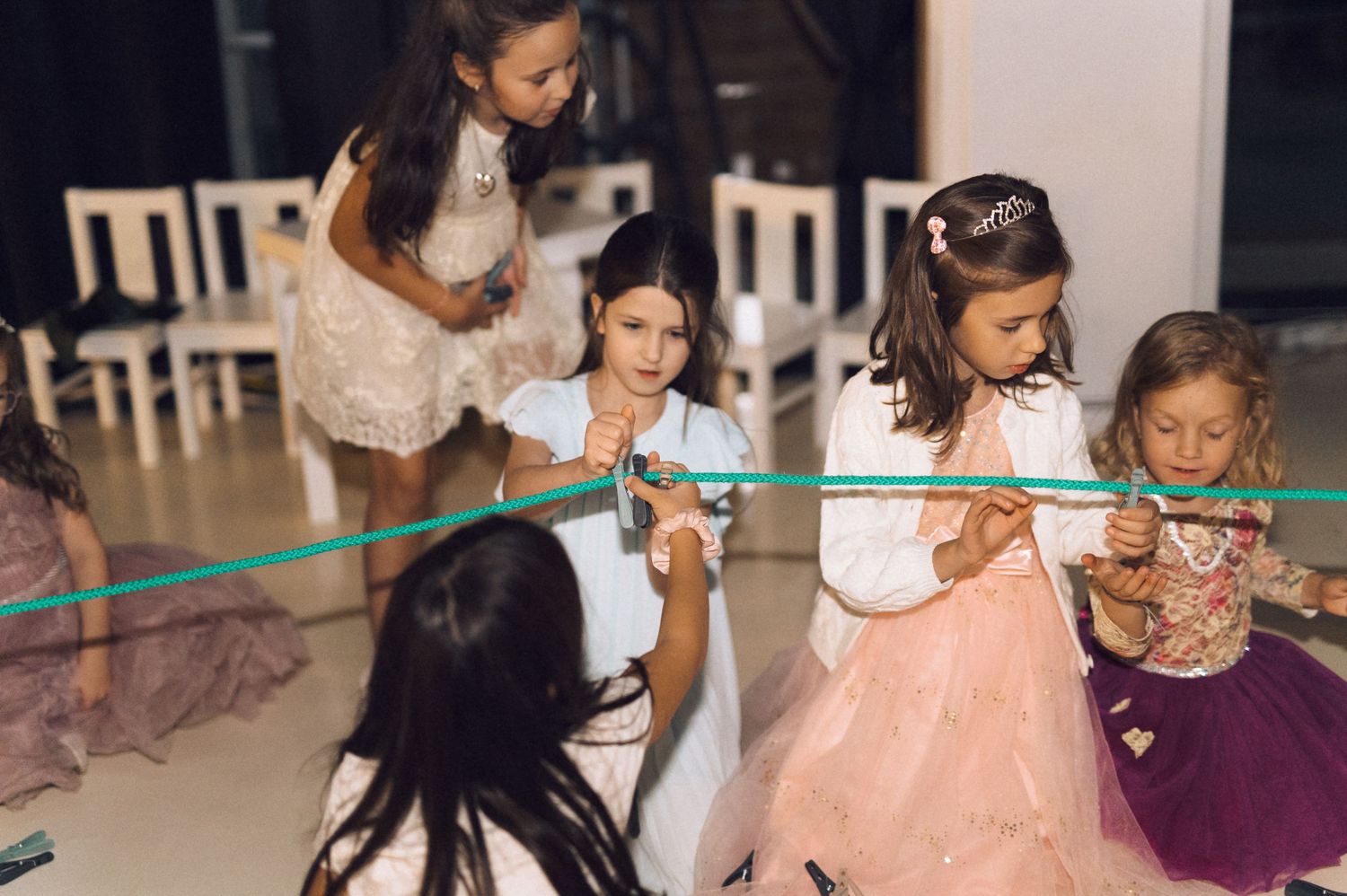 Young girls in formal dresses play a ribbon game together at an indoor celebration or party.