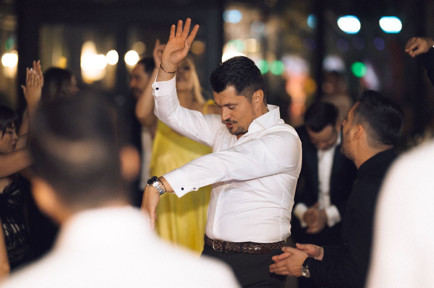 A man in a white dress shirt dances energetically with raised arms at an outdoor evening celebration.