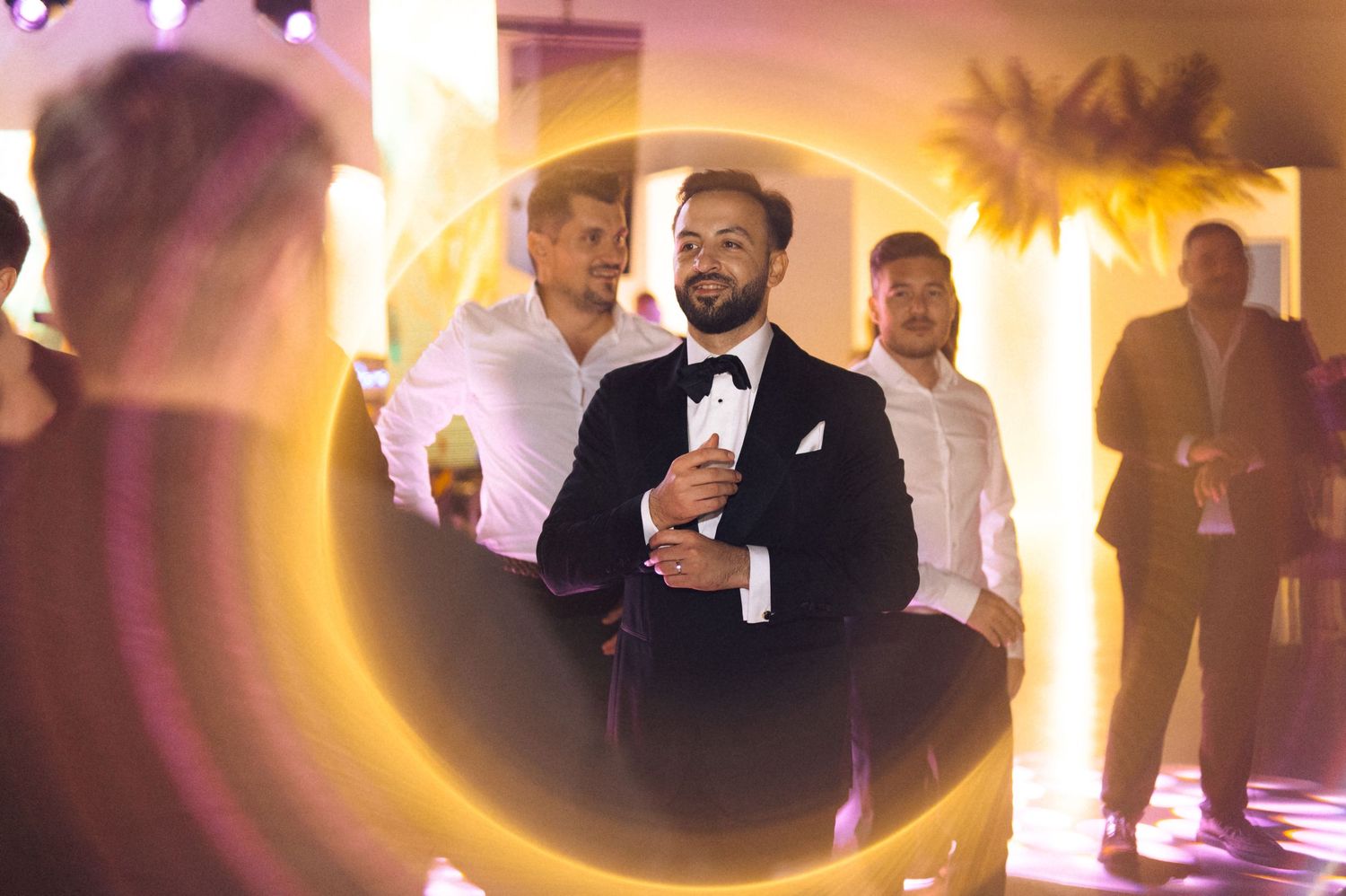 Groom in black tuxedo adjusting cufflinks surrounded by groomsmen in white shirts at elegant wedding reception.