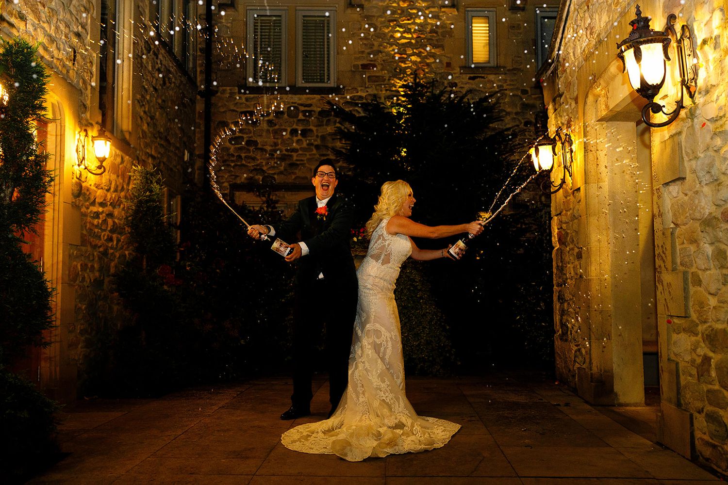 Two brides enjoying a champagne spray at night outside a Fence Gate Lodge during their wedding celebration.
