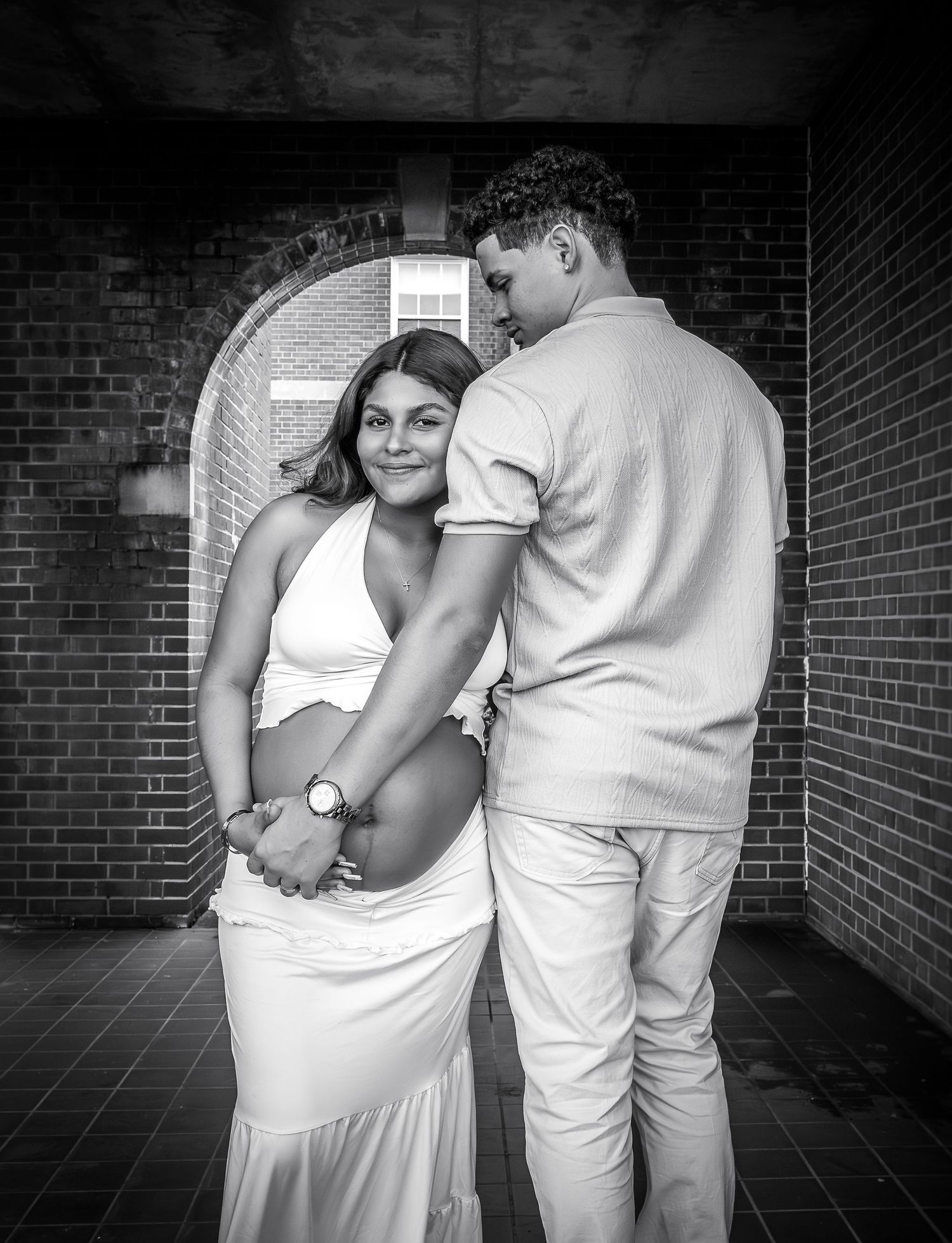 A black and white photo of a pregnant couple posing tenderly in a brick archway.
