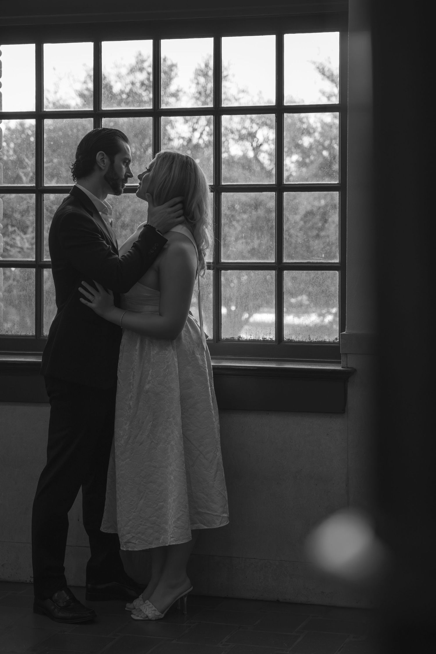 Black and white photo of a couple sharing a kiss while embracing near a large multi-pane window.