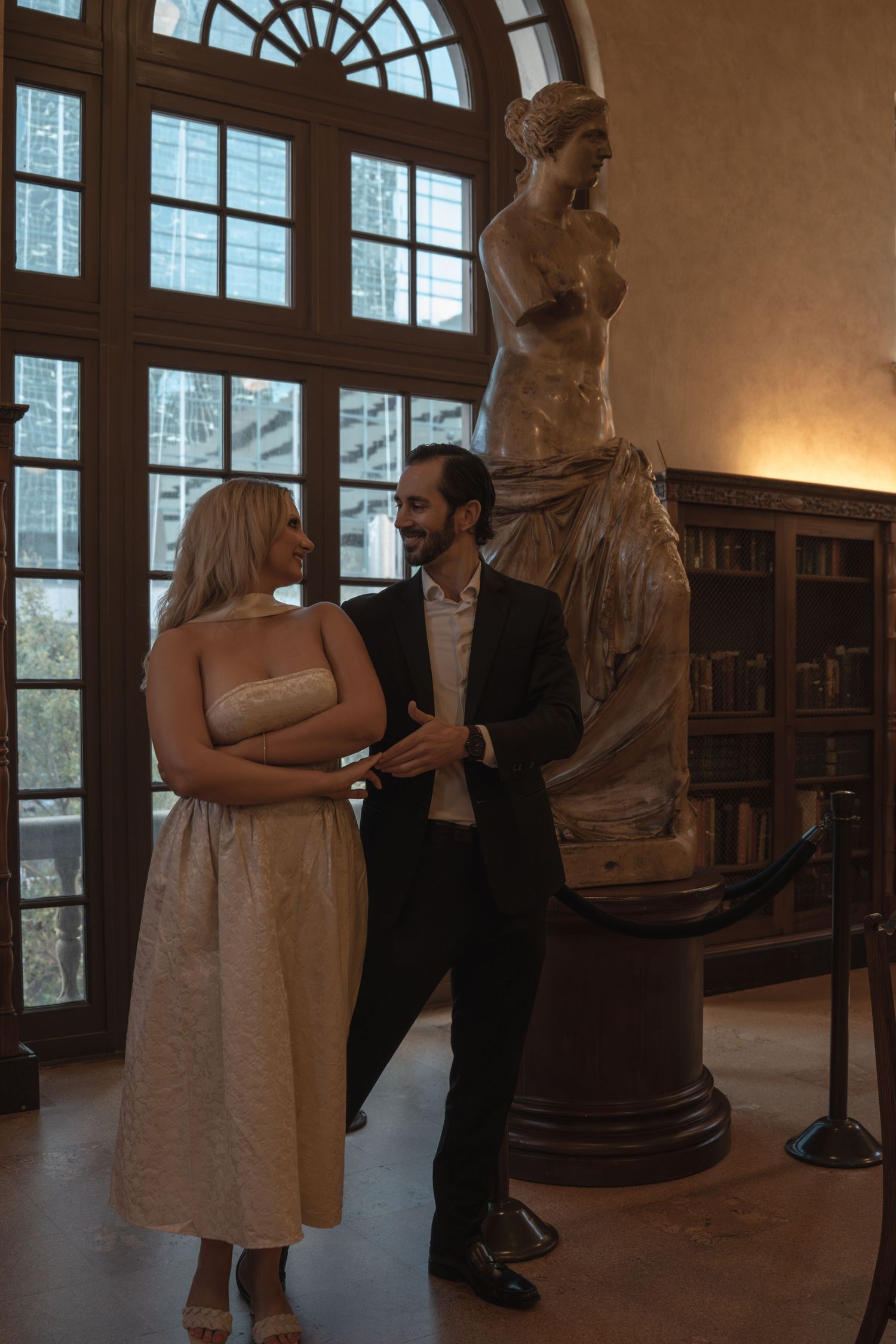 Couple smiling together beside a classical Venus de Milo statue replica in an ornate library setting.