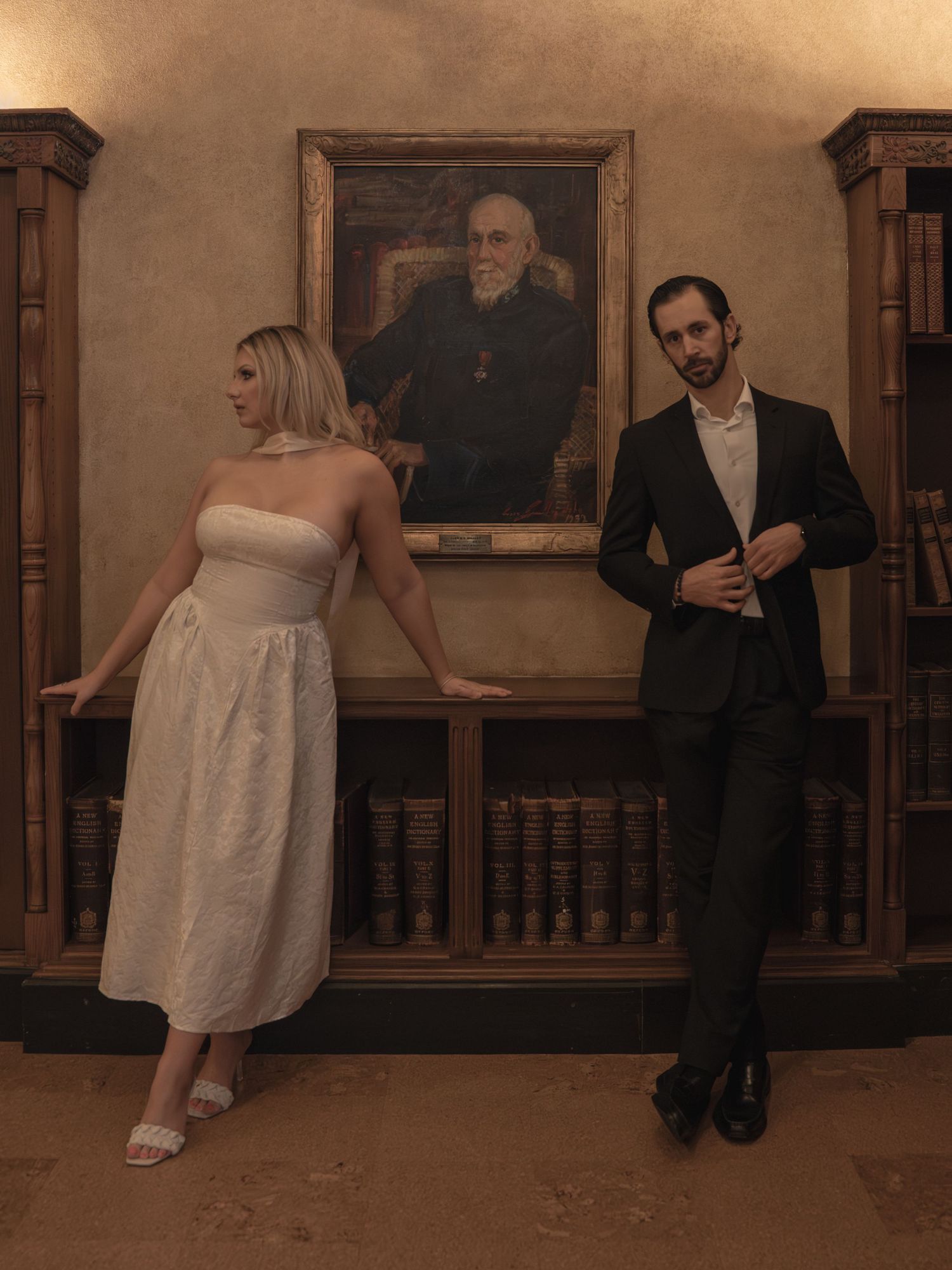 Couple in wedding attire posing casually in front of an antique portrait and bookshelf in a historic library.