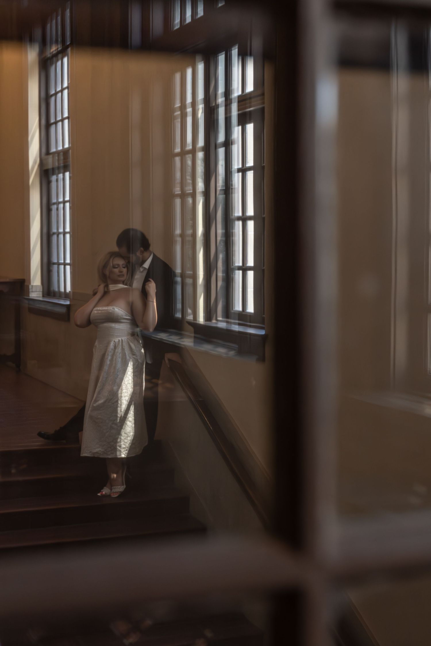 Couple sharing a tender embrace on a sunlit staircase, photographed through a decorative wooden frame.