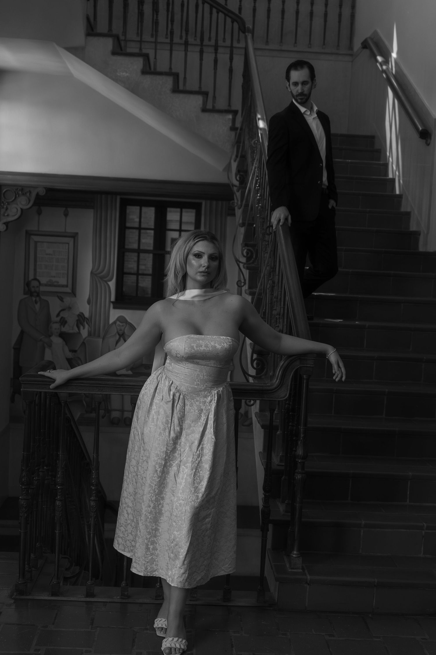 Black and white photo of a bride posing on a grand staircase with the groom descending behind her.