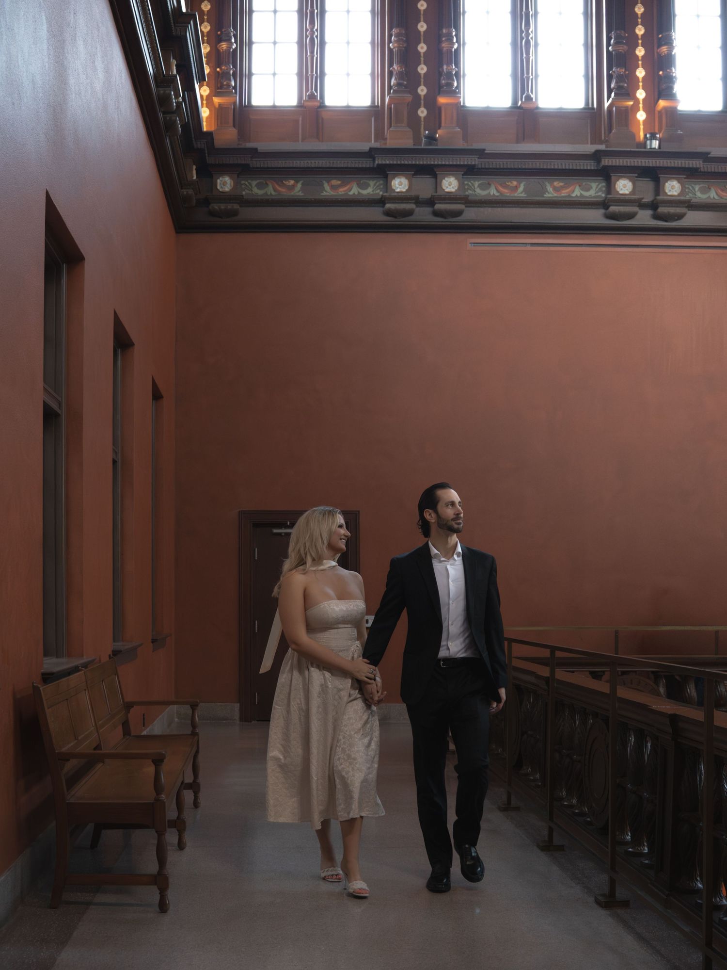 Couple holding hands walking through elegant terracotta hallway with ornate stained glass windows.