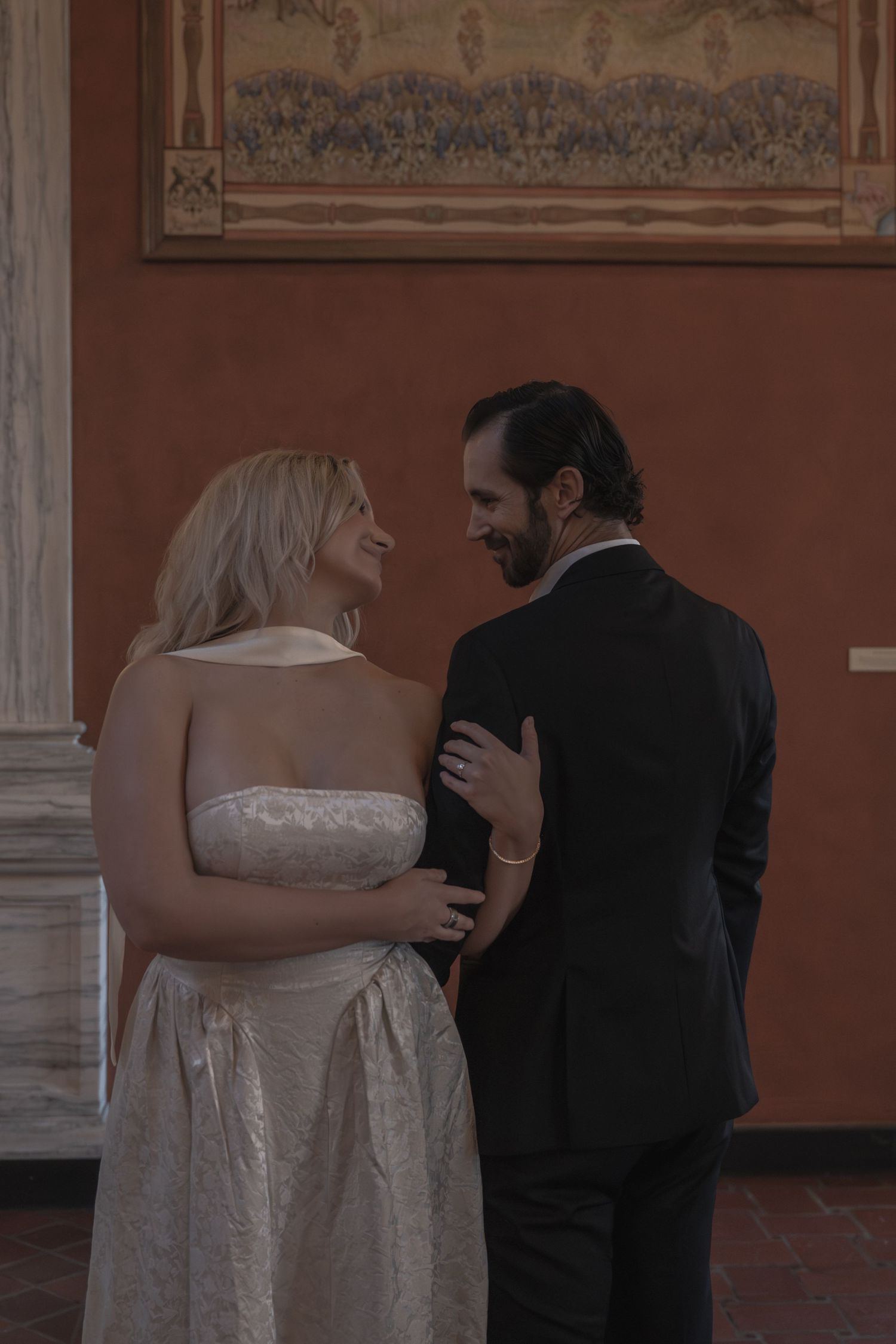 Bride in strapless white gown and groom in black suit sharing intimate moment before ornate mural.