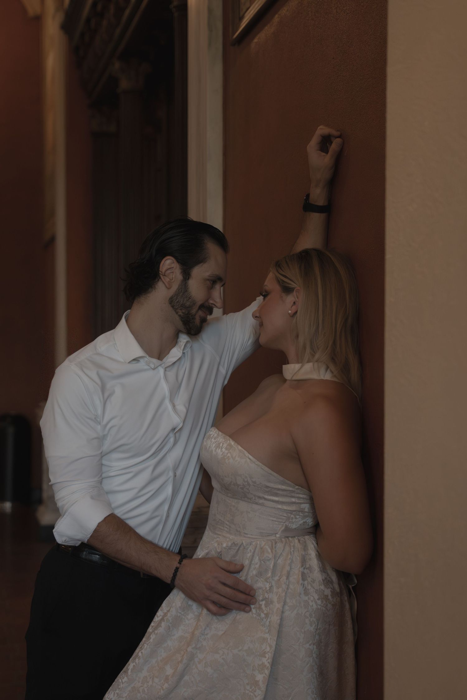 Groom in white shirt leans close to bride against terracotta wall in dimly lit corridor.