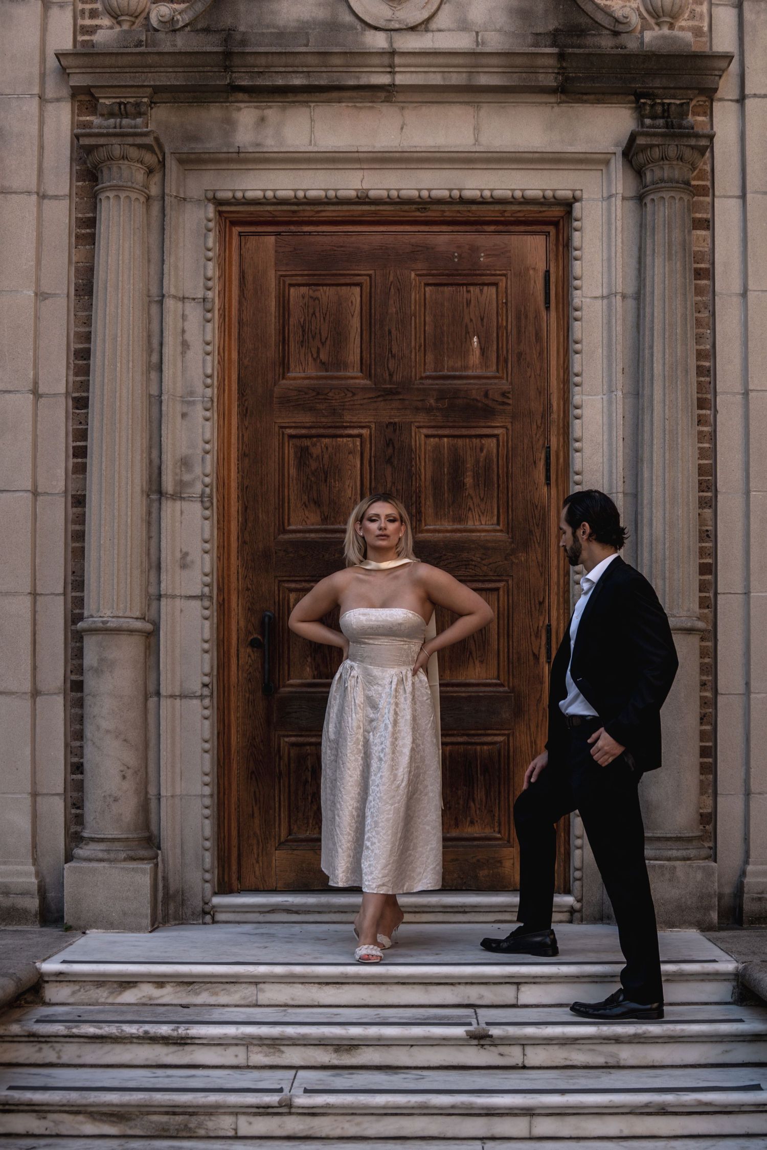 Bride in champagne midi dress poses before grand wooden door as groom admires her.