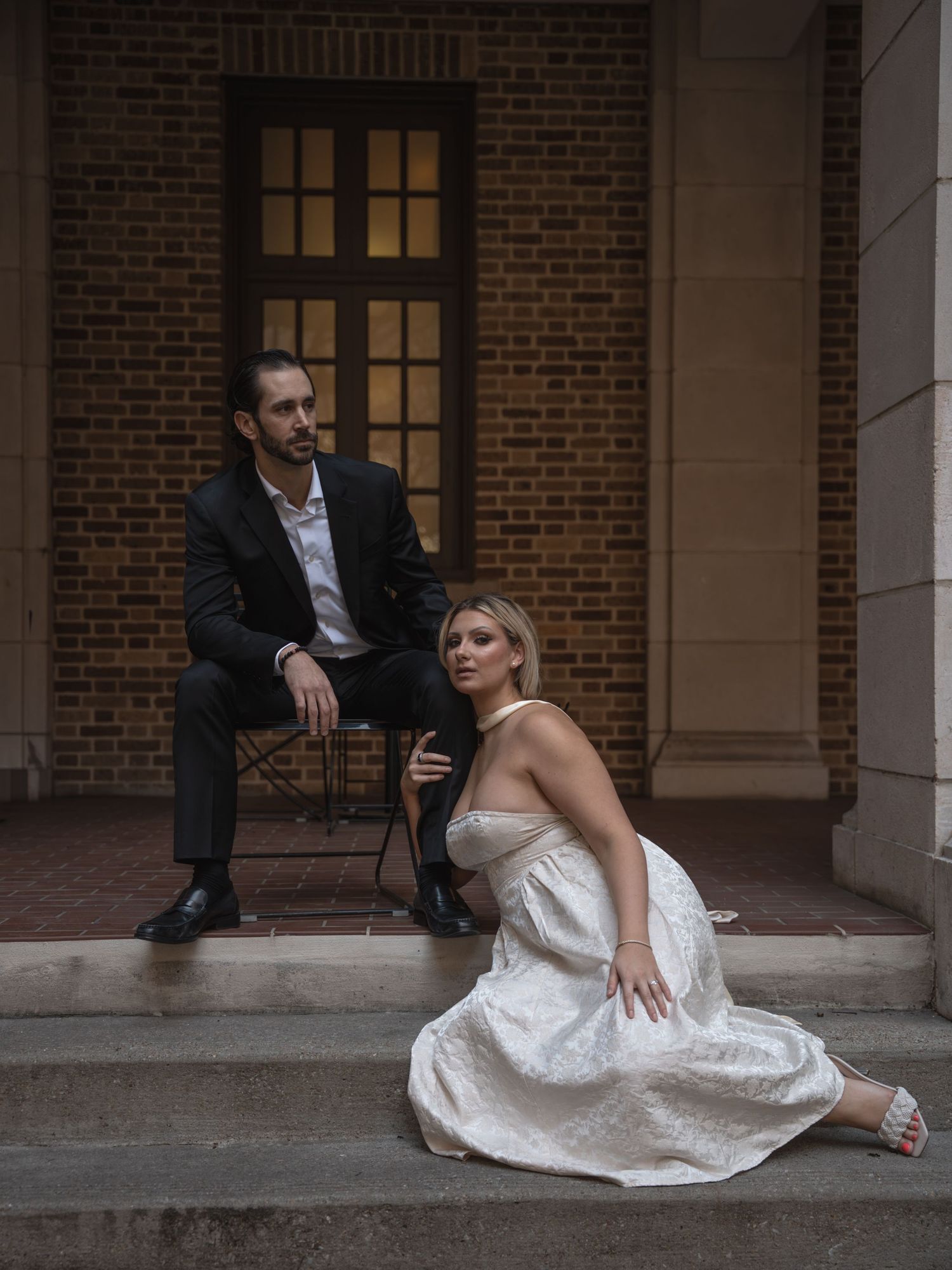 Groom seated on steps while bride sits gracefully on ground before brick building at dusk.