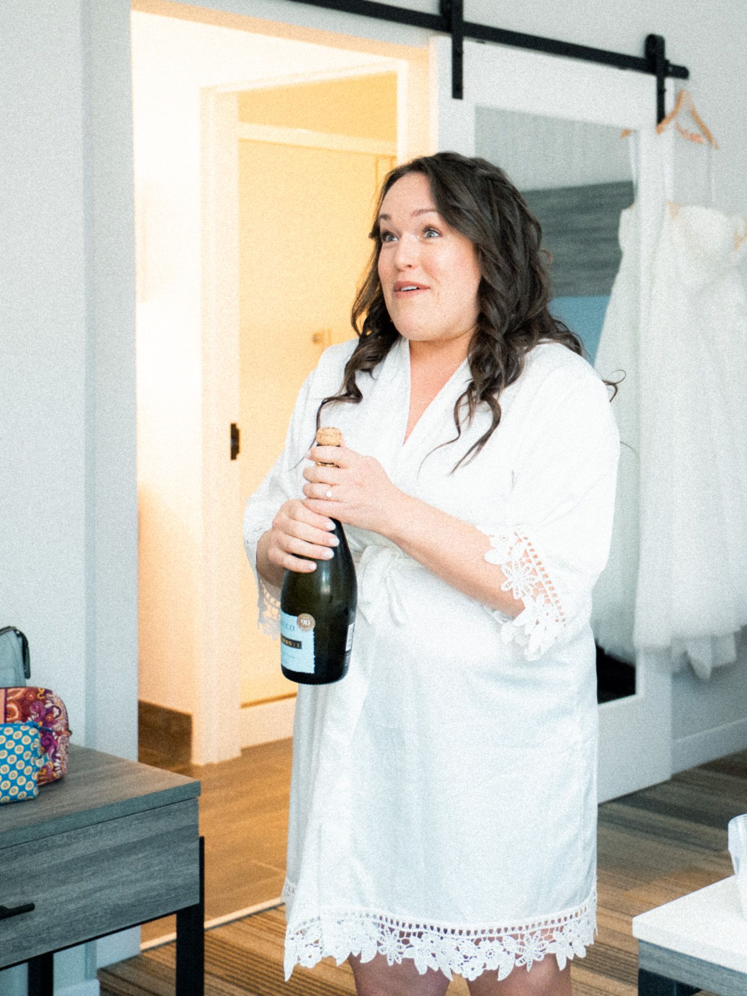 Someone in a white lace robe holds a champagne bottle while getting ready in a room with a wedding dress hanging.