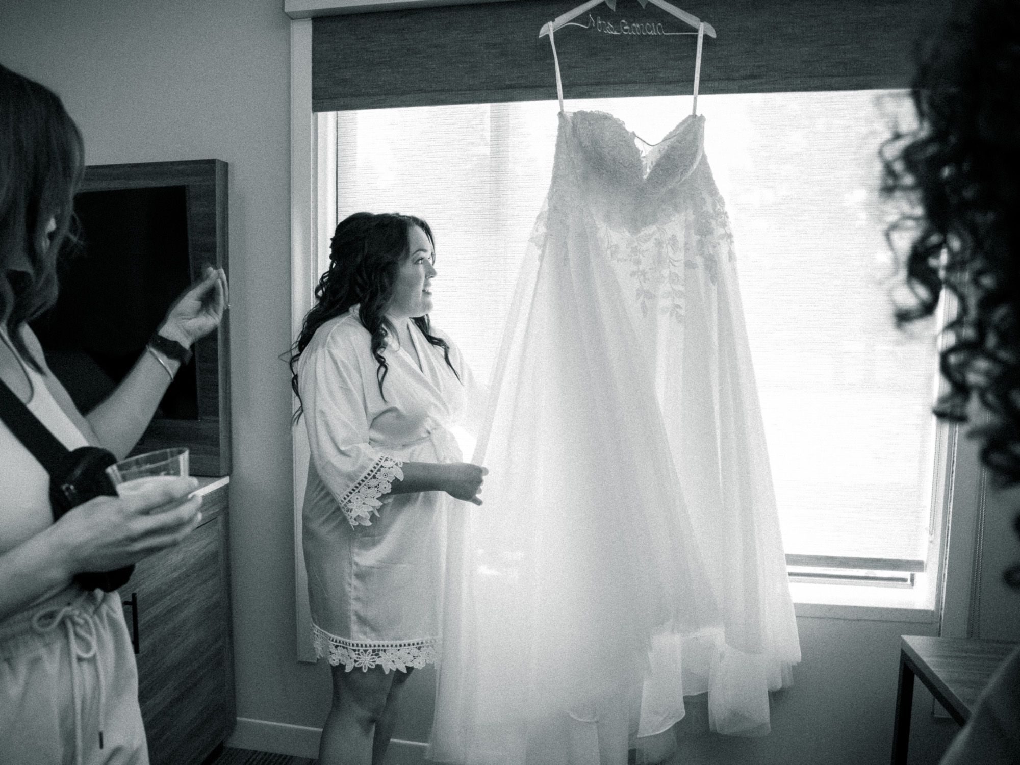 A white wedding dress hangs by a window while people in robes gather around it during wedding preparations.