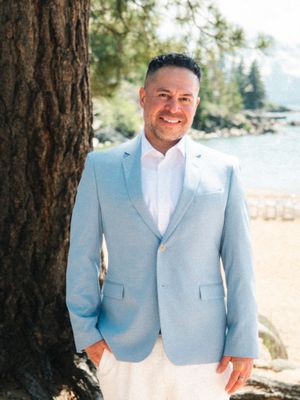 Someone wearing a light blue blazer and white shirt smiles while standing next to a tree at a lakeside beach setting.