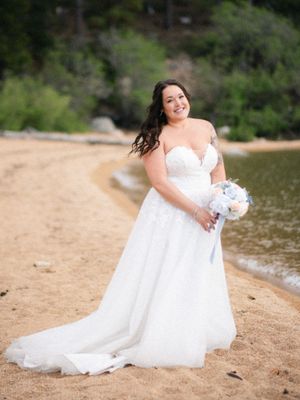 A bride in a flowing white wedding dress poses gracefully on a sandy beach by a serene lake on her special day.