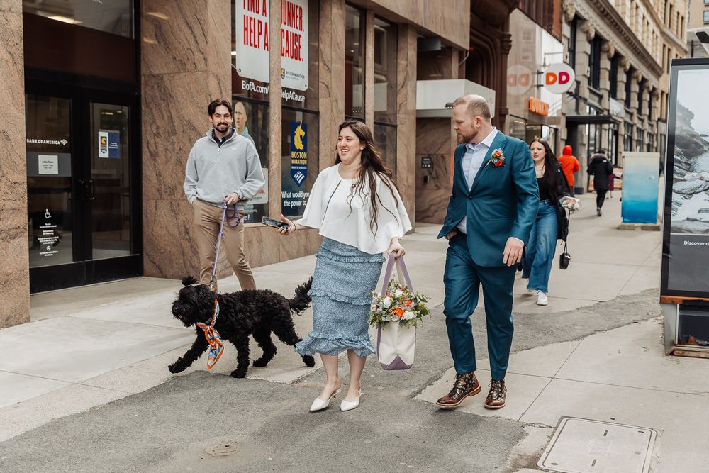 Bride and groom walking dog on city sidewalk, woman holding bouquet, man in blue suit, urban street setting.