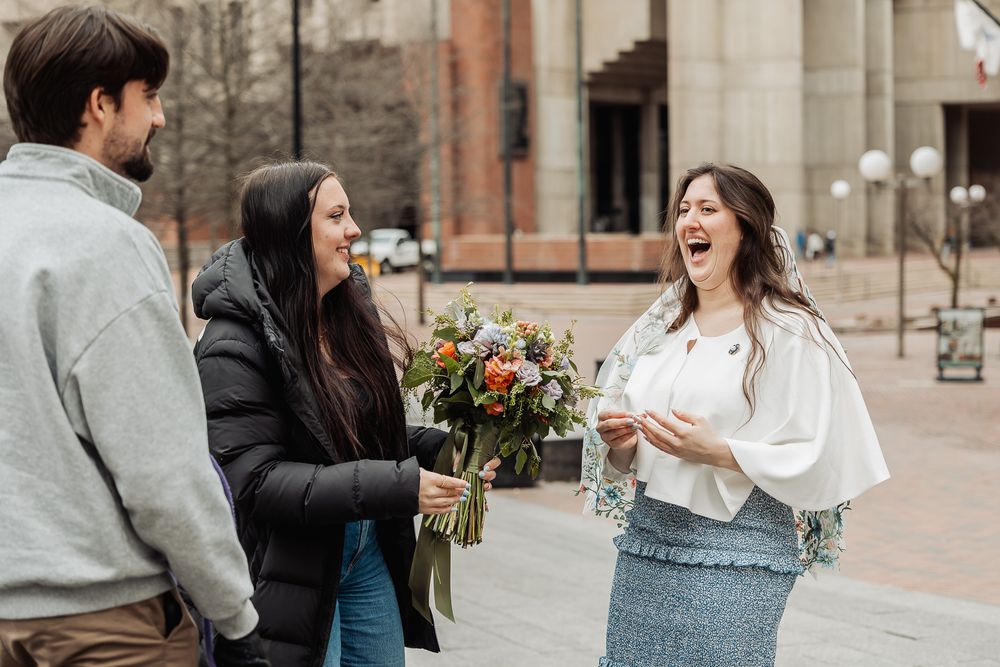 Three people laughing outdoors, woman holding colorful flower bouquet, urban building background.